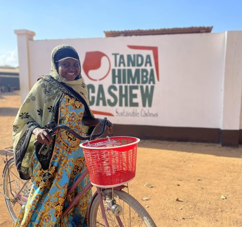 Smiling woman with bicycle in front of Tanda Himba Cashew sustainable cashew factory in Tanzania.